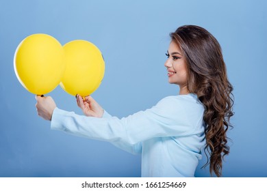 Happy Teenager Girl Is Holding Two Yellow Balloons And Smiling For Camera While Having Fun Against Isolated On Blue Background, Profile View