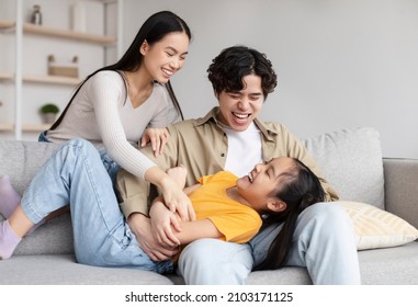 Happy Teenager Girl And Millennial Asian Man And Woman Have Fun Together, Tickling Little Daughter On Sofa In Living Room Interior. Joy, Relationship, Love And Family At Home During Covid-19 Outbreak