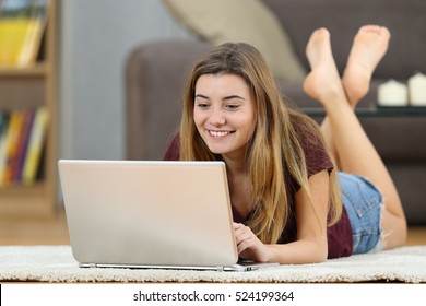 Happy teenager girl on line with a laptop lying on a carpet on the floor in the living room at home