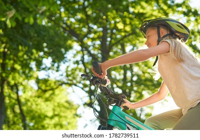 Happy Teenager Girl Rides Bike On A Summer Sunny Day In A City Park. Background Of Green Leaves Of Trees. Urban Biking. Focus On The Bicycle Handlebar