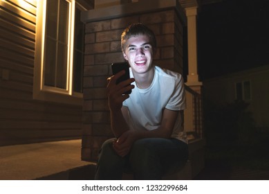 Happy Teenager On His Mobile Phone. He Is Sitting On The Front Steps Of His House At Night.