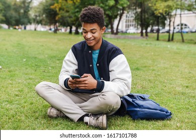 Happy Teenager Student Boy Sitting On A Lawn At The Park, Using Mobile Phone