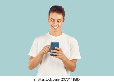 Happy teenager using modern cell phone. Studio portrait adolescent boy in T shirt standing isolated on light blue background holding smartphone, scrolling news feed, looking at funny memes and smiling - Powered by Shutterstock