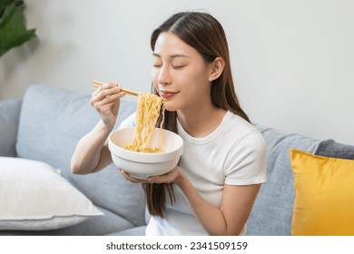 Happy temptation, cute asian young student woman, girl using chopsticks eating instant ramen, noodles soup in bowl while watching TV in living room at home, cooking meal fast food lifestyle of person.
