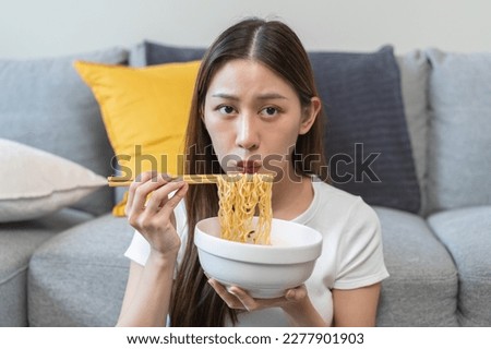 Happy temptation, cute asian young student woman, girl using chopsticks eating instant ramen, noodles soup in bowl while watching TV in living room at home, cooking meal fast food lifestyle of person.
