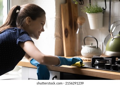 Happy tenant girl using blue protective rubber gloves, rug, cleaning kitchen countertop, cooktop, smiling, enjoying cleanup. Household chores, cleaning service, domestic hygiene concept
