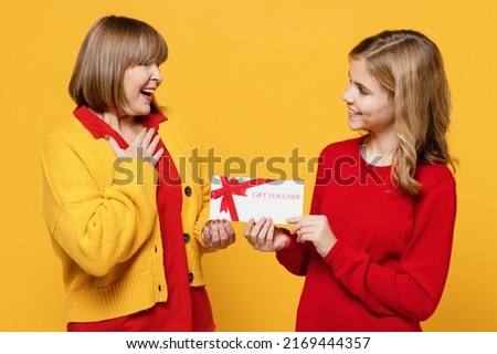 Happy thankful woman 50s in red shirt have fun with teenager girl 12-13 years old Grandmother granddaughter hold give gift certificate coupon voucher card for store isolated on plain yellow background