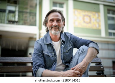 Happy thinking of life middle aged man leaned his hands on knee looking down his head sitting on the bench in old town. Happy mature grey bearded man with positive emotions on his face. - Shutterstock ID 2180454513
