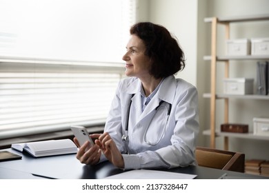 Happy thoughtful mature doctor woman using online app on mobile phone, consulting internet, texting message on chat, making appointment with patients, looking away at window, thinking at work table - Powered by Shutterstock