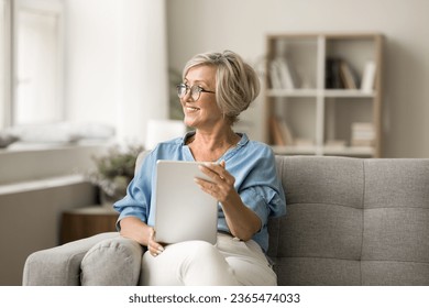 Happy thoughtful mature old woman using digital device at home, holding tablet computer on comfortable sofa, looking away with good thoughts, inspiration, smiling, thinking, daydreaming - Powered by Shutterstock