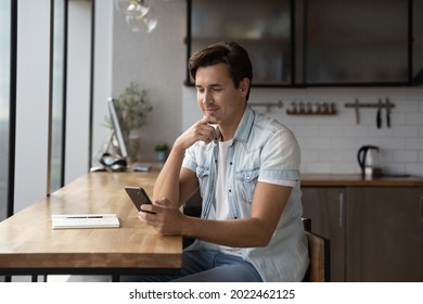 Happy Thoughtful Millennial Guy Using App, Online Service On Smartphone At Home Workplace Table, Looking At Screen, Browsing Internet, Reading Text, Typing Message. Man Making Call On Mobile Phone