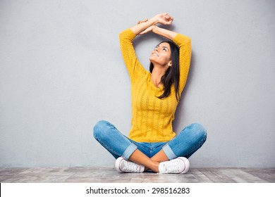 Happy thoughtful woman sitting on the floor with crossed legs and looking up over gray background