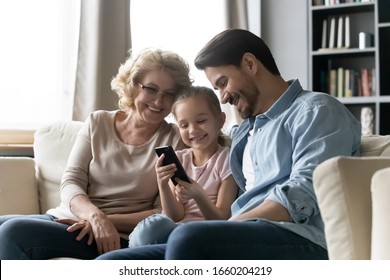 Happy three family generations sit rest on couch in living room watch funny video on smartphone together, smiling little girl with young father and grandmother using cellphone gadget at home
