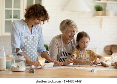 Happy three generations of Caucasian women have fun cooking together bread or pie in home kitchen. Cute little 9s girl child baking with mature 60s grandmother and young mother, prepare dessert.