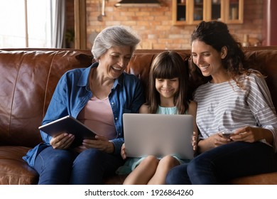 Happy three generations of Latino women sit relax on couch at home using modern electronic gadgets together. Smiling little Hispanic girl with mom and grandmother rest indoors have fun with devices. - Powered by Shutterstock