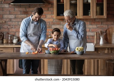 Happy Three Generations Of Men Wearing Aprons Cooking Standing In Kitchen Together, Mature Grandfather In Glasses With Grownup Son And 8s Grandson Cutting Fresh Vegetables, Enjoying Leisure Time