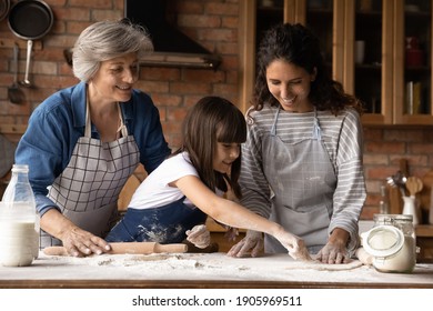 Happy three generations of women cooking handmade pastry, kneading, rolling dough, standing in kitchen, happy little girl with smiling mother and mature grandmother enjoying leisure time at home