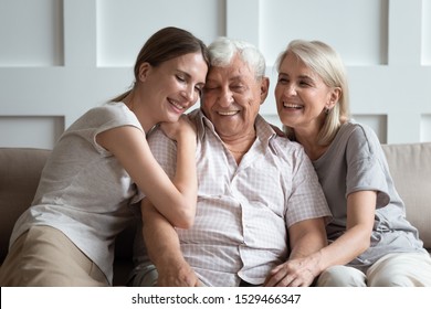 Happy three-generation family hugging sit on sofa enjoy time together at home, laughing grown up daughter with 70s dad and middle-aged 50s mom warm relations, intergenerational relative people concept