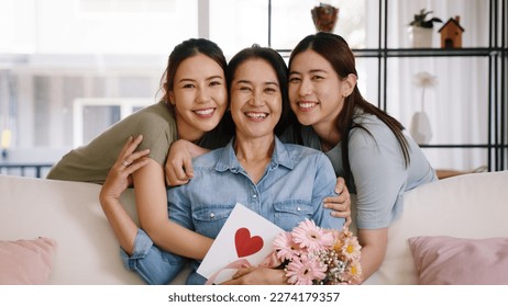 Happy time Mother day grown up child looking at camera cuddle hug give flower gift box red heart card to mature mum. Love kiss care mom asia middle age adult people smile enjoy sitting at home sofa. - Shutterstock ID 2274179357