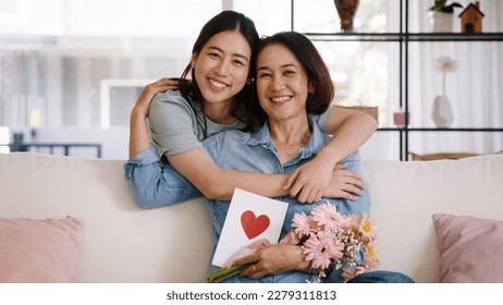 Happy time Mother day grown up child looking at camera cuddle hug give flower gift box red heart card to mature mum. Love kiss care mom asia middle age adult people smile enjoy sitting at home sofa. - Powered by Shutterstock