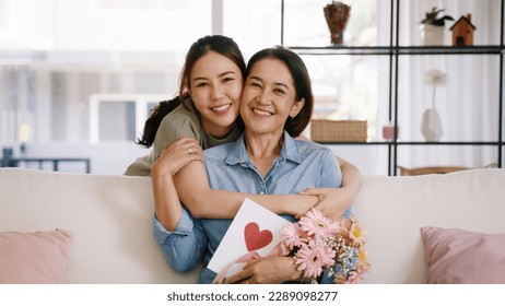 Happy time Mother day grown up child looking at camera cuddle hug give flower gift box red heart card to mature mum. Love kiss care mom asia middle age adult people smile enjoy sitting at home sofa. - Shutterstock ID 2289098277