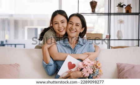 Happy time Mother day grown up child looking at camera cuddle hug give flower gift box red heart card to mature mum. Love kiss care mom asia middle age adult people smile enjoy sitting at home sofa.