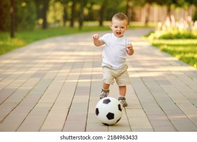 Happy Toddler Baby Boy Is Playing Walking Behind A Soccer Ball On A Stone Path, First Steps. Smiling Child In White Clothes Walks With A Ball, Age One Year