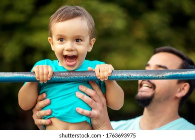 Happy Toddler Boy Doing Pull Ups Exercise, Child Smiling Happily Having Pulled Up On Steel Bar Outdoors In Summer Day. Cheerful Father Supporting Kid. Success, Achievement And Healthy Family Concept