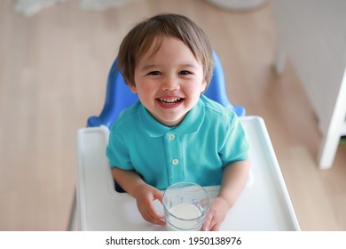 Happy Toddler Boy Drinking White Milk On High Chair At Home. Mixed Race Asian-German Child Holding A Glass Of Drink With Laughing Face. Kid With Healthy Nutrition.