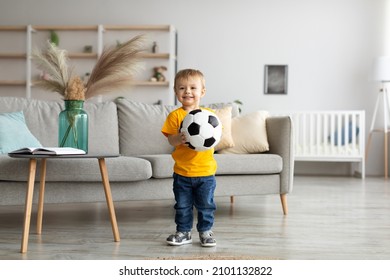 Happy Toddler Boy Football Fan Posing With Soccer Ball And Smiling At Camera, Having Fun At Home In Living Room Interior, Copy Space. Sports Activities And Games For Kids