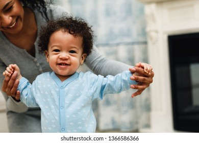 Happy Toddler Boy Learning To Walk. Mother Assisting Son In Walking At Home.