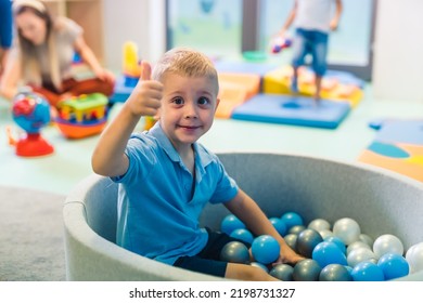 Happy Toddler Boy Playing In A Ball Pit Full Of Colorful Balls, Showing Thumb Up. Sensory Play At The Nursery School For Kids Wellness. High Quality Photo