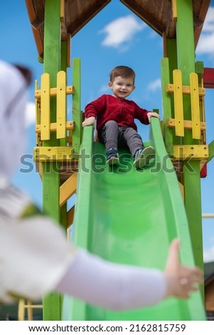 Happy toddler boy playing on a slide at a playground with her mother.
