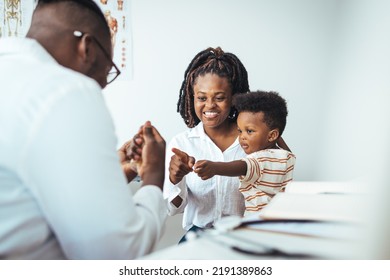 A Happy Toddler Boy Sits In His Mother' Lap At The Pediatrician. He Plays Patacake With A Doctor. Doctor Giving Prescription To African American Mother With Baby Son At Clinic. 