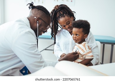 A Happy Toddler Boy Sits In His Mother' Lap At The Pediatrician. He Plays Patacake With A Doctor. Doctor Giving Prescription To African American Mother With Baby Son At Clinic. 