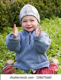 Happy Toddler Child Sitting In Grass In A Garden And Clapping His Hands. 