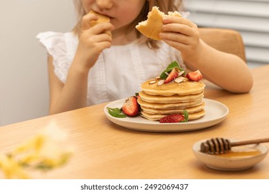 Happy toddler girl eats delicious pancakes for breakfast at table closeup. Little child bites round crepe with berries and honey at home. Handmade meal - Powered by Shutterstock