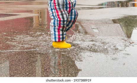 Happy Toddler Running And Jumping Into Pools Of Water In The Rain, Wearing Yellow Rubber Boots. Carefree Kid With Colorful Rain Boot And Suit Splashing Water In An Urban Scenery With Reflections