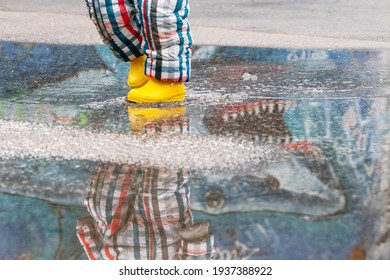 Happy Toddler Running And Jumping Into Pools Of Water In The Rain, Wearing Yellow Rubber Boots. Carefree Kid With Colorful Rain Boot And Suit Splashing Water In An Urban Scenery With Reflections