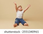 Happy toddler in white t-shirt and jeans sits against beige studio background, reaching upwards with big smile, expressing joy and curiosity. Concept of parenthood, natural beauty, love and care. Ad.