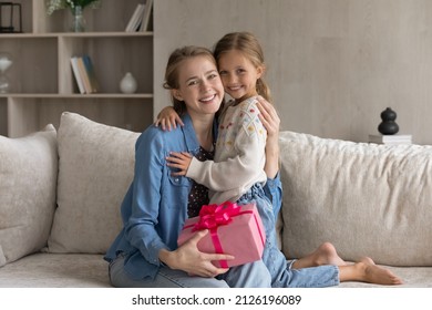 Happy Touched Young Mom And Cheerful Cute Daughter Kid Celebrating Mothers Day, Birthday, 8 March, Holding Pink Gift Wrap, Hugging On Couch With Love, Tenderness Looking At Camera. Family Portrait