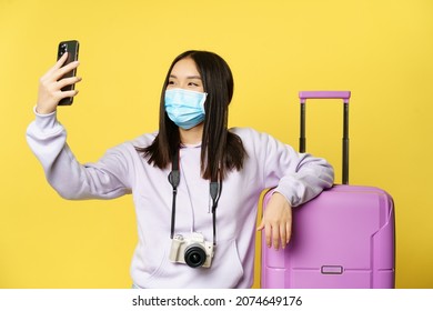 Happy tourist, asian girl on vacation taking selfie in medical mask near suitcase, photographing on smartphone, yellow background