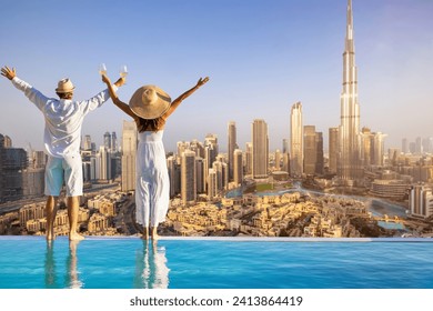A happy tourist couple on vacation time stands by the pool edge and enjoys the panoramic sunset view of the Dubai city skyline, UAE