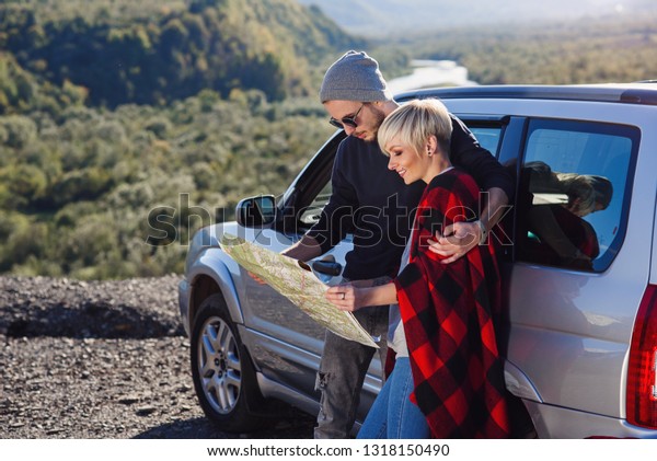 Happy tourist couple with paper map near\
rented car. Trendy young people using map. Traveling by car on\
summer vacations in mountains. Hipster man and woman having trip\
outdoors. Landscape\
background.