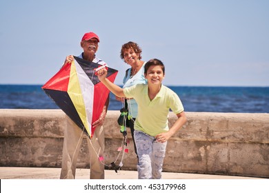 Happy Tourist Family On Holidays In Cuba. Hispanic Grandpa, Grandma And Grandson Having Fun Near The Sea. Boy And Grandparents Running And Playing With Kite 