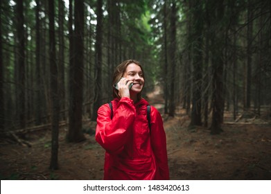 Happy tourist girl talking on the phone on a hike, standing in the woods, wearing a red jacket and backpack, looking away and smiling. Portrait of positive tourist woman hiking, talking on the phone. - Powered by Shutterstock