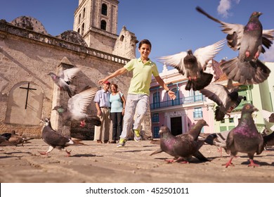 Happy Tourist On Holidays During Vacation Trip. Hispanic People Traveling In Havana, Cuba. Grandpa And Grandson Feeding Birds, With Child Running Chasing Pigeons