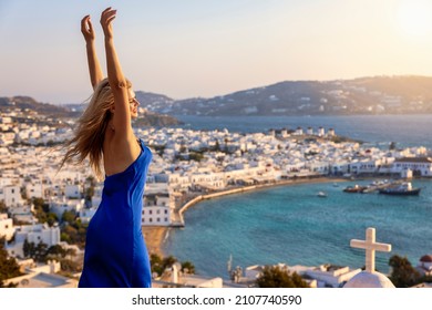 A Happy Tourist Woman In A Blue Party Dress Enjoys The Elevated Sunset View To The Town Of Mykonos Island, Greece