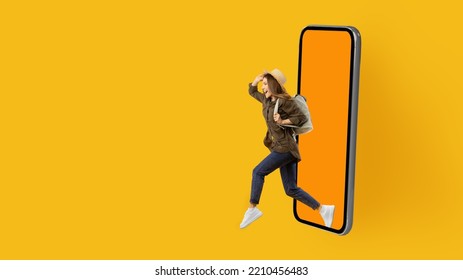 Happy Tourist Woman Running Out Of Large Mobile Phone Screen Advertising Great Travel Offer Posing With Backpack Over Yellow Studio Background. Touristic Application. Panorama, Copy Space