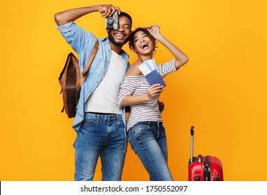 Happy Tourists. Excited African Couple Ready For Vacation, Posing With Suitcase, Tickets And Photo Camera Over Yellow Studio Background.